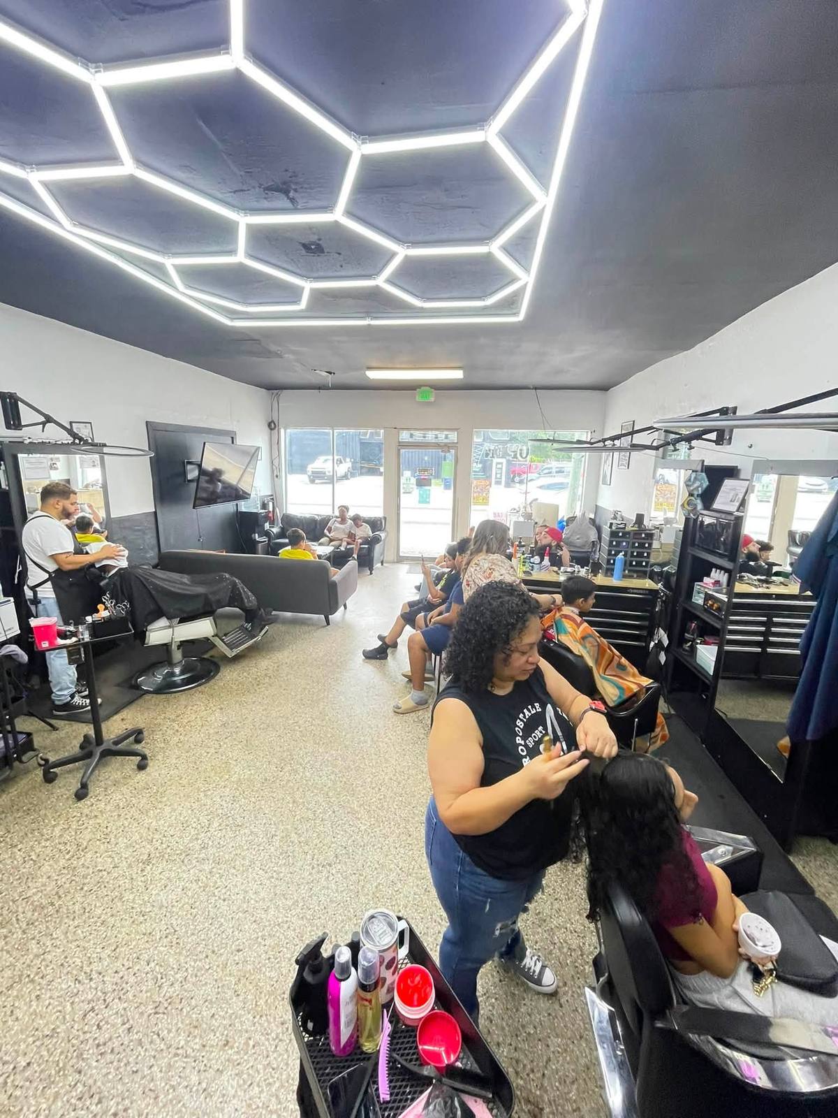 The shop floor from the door: a hexagonal LED tube grid across the black ceiling, stations with black tool chests and framed mirrors on ring-light arms, barbers working and families waiting.