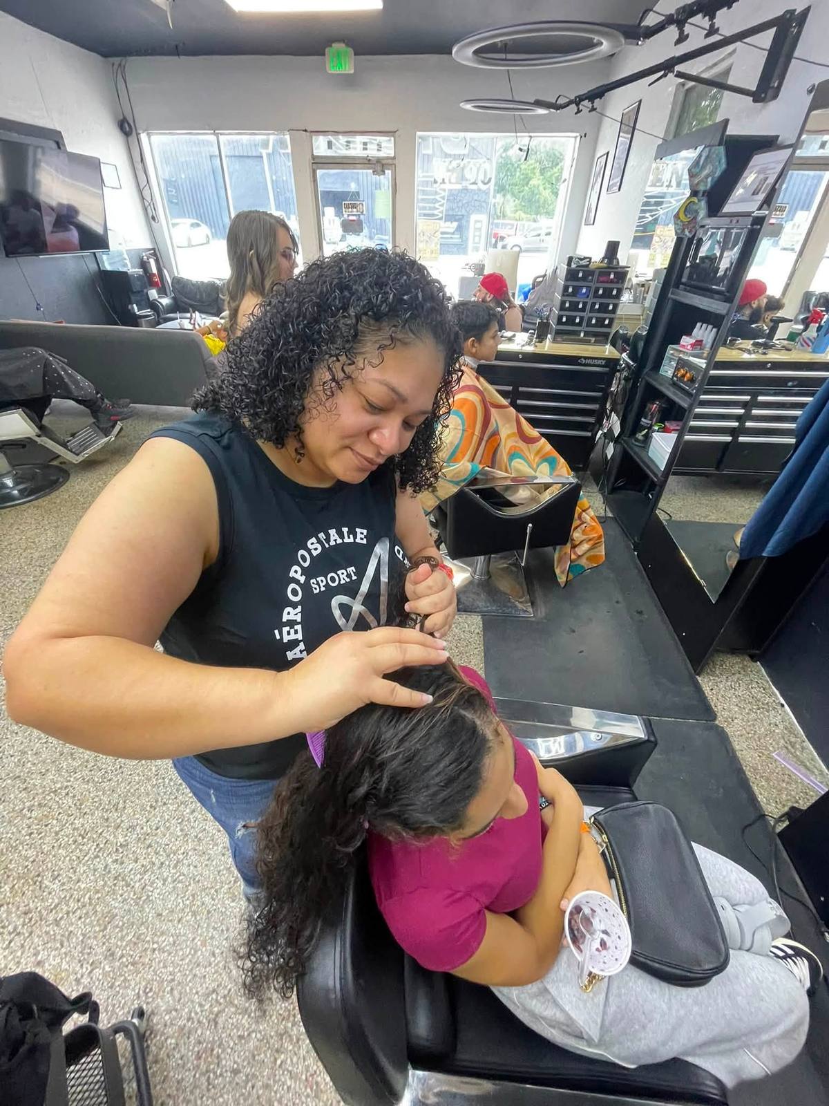 A stylist sectioning and styling a seated client&rsquo;s long curly hair in the shop &mdash; the room does women&rsquo;s styling, not barbering only.