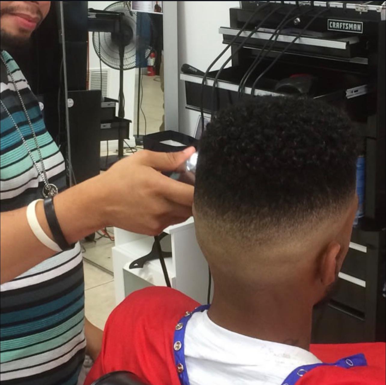 Barber&rsquo;s hands running clippers through the back of a client&rsquo;s mid fade, station and tool chest behind.