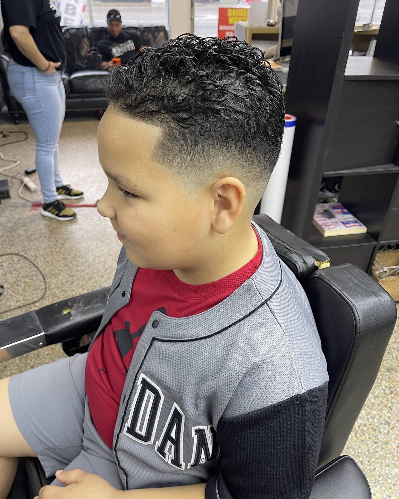 Boy in the barber chair at Refined Barber Lounge, finished high taper fade with the curly natural top left long. Leather waiting sofas and the N. Nebraska Ave storefront window behind him.